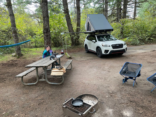 Woman seated at a picnic table in a camping spot in the forest with an SUV with a rooftop tent.