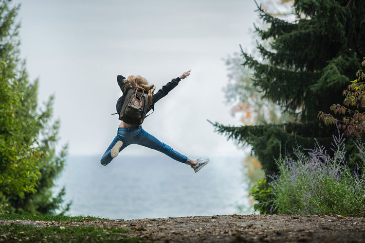 Woman jumping in the air wearing a backpack on a trail with pathway and trees.