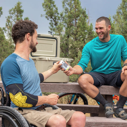 a man in a wheelchair and a man sitting on a bench cheers their beers with a cooler behind them.